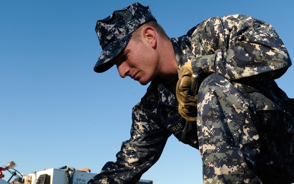A U.S. Navy sailor in Navy Working Uniform (NWU) Type I digital camouflage is crouching down on what appears to be a flight deck or tarmac, performing a hands-on maintenance or inspection task. The sailor is wearing gold-colored work gloves and black combat boots. A white ground support vehicle is visible in the background on the airfield.
