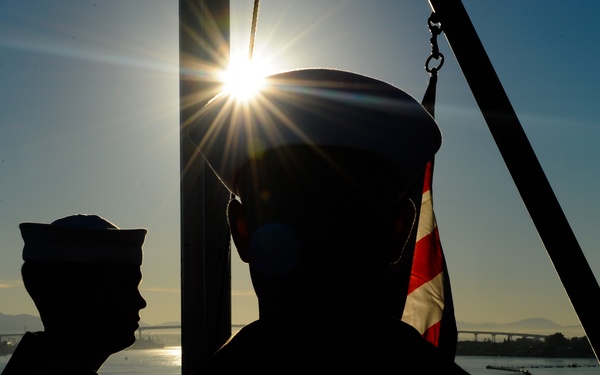 Two military personnel in silhouette are photographed against a sunrise or sunset over a body of water, likely during a flag ceremony aboard a naval vessel. A partially visible red and white flag — consistent with a U.S. Navy jack or national ensign — is being handled near a flagpole or mast. A bridge and distant shoreline are visible in the background.