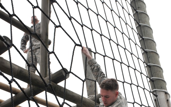 A U.S. Army soldier in ACU (Army Combat Uniform) pattern camouflage is climbing a large rope net obstacle on a military training course. A second soldier in uniform is visible in the upper background, standing on a wooden platform structure. The setting appears to be an outdoor obstacle course during overcast, cold-weather conditions.