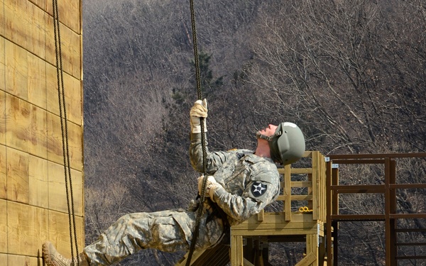 Two U.S. Army soldiers in ACU (Army Combat Uniform) camouflage are conducting rope-climbing training on a vertical wooden wall at a military obstacle course. The upper soldier is actively climbing the wall using ropes while wearing a helmet, and a unit patch (2nd Infantry Division) is visible on their shoulder. A second soldier at the base appears to be beginning their climb or providing support, with wooden training structures and a barbed-wire perimeter fence visible in the background.
