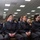 A group of young individuals wearing matching dark gray uniforms with shoulder epaulettes and light blue collar accents are seated in rows in what appears to be a formal meeting room or auditorium. Four uniformed individuals are prominently visible in the foreground, with additional civilians seated behind them. The setting suggests a formal ceremony, briefing, or official gathering, likely at a law enforcement or military academy.