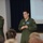 Two U.S. Air Force personnel in olive green flight suits are standing at the front of a briefing room or auditorium. The individual in the foreground appears to be speaking or presenting, while the second stands with arms crossed in the background. A projection screen displaying what appears to be aircraft imagery is visible behind them.