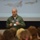 A smiling military aviator in an olive green flight suit is giving a presentation to an audience, likely children or students. He is standing in front of a projected slideshow displaying images of military aircraft, including what appears to be an F-15 and a P-51 Mustang. He is giving a thumbs-up gesture and wearing a name tag and unit patch.