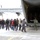 A group of civilians, likely students or visitors, are being guided toward the open rear cargo ramp of a large military transport aircraft on a snow-dusted airfield. A crew member in a flight suit is directing the group as they approach the aircraft. Several uniformed military personnel are visible standing inside the cargo bay, apparently receiving the visitors.