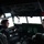 Three young men are inside a large aircraft cockpit, which appears to be a military or transport aircraft based on the complexity of the instrument panel. The central figure is seated in what appears to be the pilot's seat, holding the control yoke, while the other two observe. The cockpit features multiple green-lit digital displays, overhead panels with switches, and dual control columns.