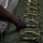 A person wearing military camouflage uniform and black tactical gloves is inspecting or arranging a row of olive-drab parachute packs lined up in sequence. Each pack is fitted with a large silver snap hook (carabiner-style connector) attached to yellow static line webbing with blue safety indicators. The image appears to depict pre-jump equipment inspection or rigging preparation in a military airborne context.