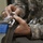 A person wearing US Army ACU (Army Combat Uniform) digital camouflage is handling parachute rigging equipment. Their hands are manipulating a metal carabiner or snap hook attached to a cable loop, connected to olive drab webbing with a distinctive blue strap. The close-up shot focuses on the hands and equipment, suggesting parachute inspection or rigging activity.