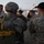 A group of military personnel are gathered on a tarmac or airfield, apparently preparing for or debriefing after a parachute operation. At least one soldier is visibly wearing a parachute pack with yellow static lines/straps and a combat helmet in ACU (Army Combat Uniform) pattern. A military transport aircraft, likely a C-130 or similar, is visible in the background.