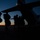 A silhouetted group of military personnel carrying full combat gear and backpacks are boarding a large military transport aircraft at dusk or dawn. The scene is captured in dramatic low-light conditions with a deep orange and blue twilight horizon in the background. A single light is visible on the aircraft, and the tarmac surface is faintly discernible beneath the figures.