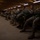 A group of military personnel in U.S. Army ACU (Army Combat Uniform) camouflage are seated on wooden benches inside a large wooden building, appearing to be a staging or holding area. The soldiers are equipped with parachute rigs and large rucksacks, suggesting they are preparing for an airborne operation. The facility appears to be a military 'chalk house' or airborne staging facility, likely at a U.S. Army installation.