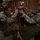 Military personnel in US Army ACU (Army Combat Uniform) camouflage are inside a wooden building, preparing parachute equipment before an airborne operation. Two soldiers in the foreground are actively inspecting or adjusting parachute gear on a table, while additional soldiers with parachute packs are visible in the background. The setting appears to be a pre-jump staging area or rigging shed.