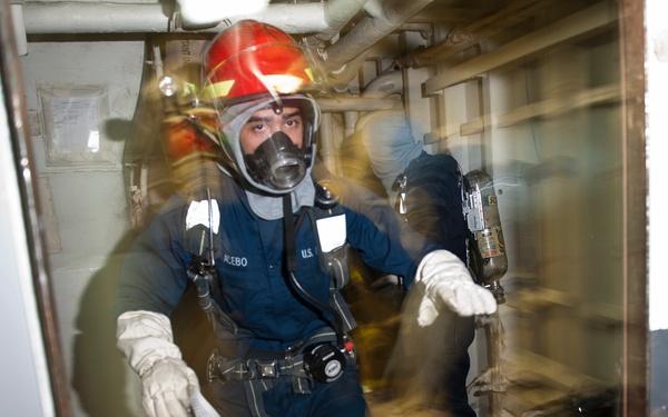 A person identified by a name tag reading 'ACEBO' wearing U.S. military or Coast Guard uniform is equipped with full firefighting/damage control gear including a red helmet, full-face respirator/gas mask, SCBA (self-contained breathing apparatus), white gloves, and a utility belt. The individual appears to be navigating through a narrow interior corridor of a ship or vessel during what appears to be a damage control or firefighting drill. The image is photographed through a glass or transparent surface, creating distortion around the edges.
