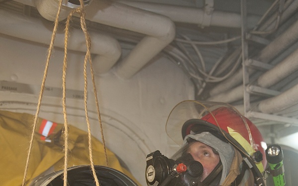 A person wearing full firefighting or damage control gear, including a red helmet, Scott brand SCBA (Self-Contained Breathing Apparatus) face mask, and white protective gloves, is working with a large circular duct or pipe opening suspended by rope. The setting appears to be the interior of a naval vessel, indicated by the overhead piping, cables, and industrial infrastructure visible in the background. The individual appears to be conducting damage control or emergency response training or operations.