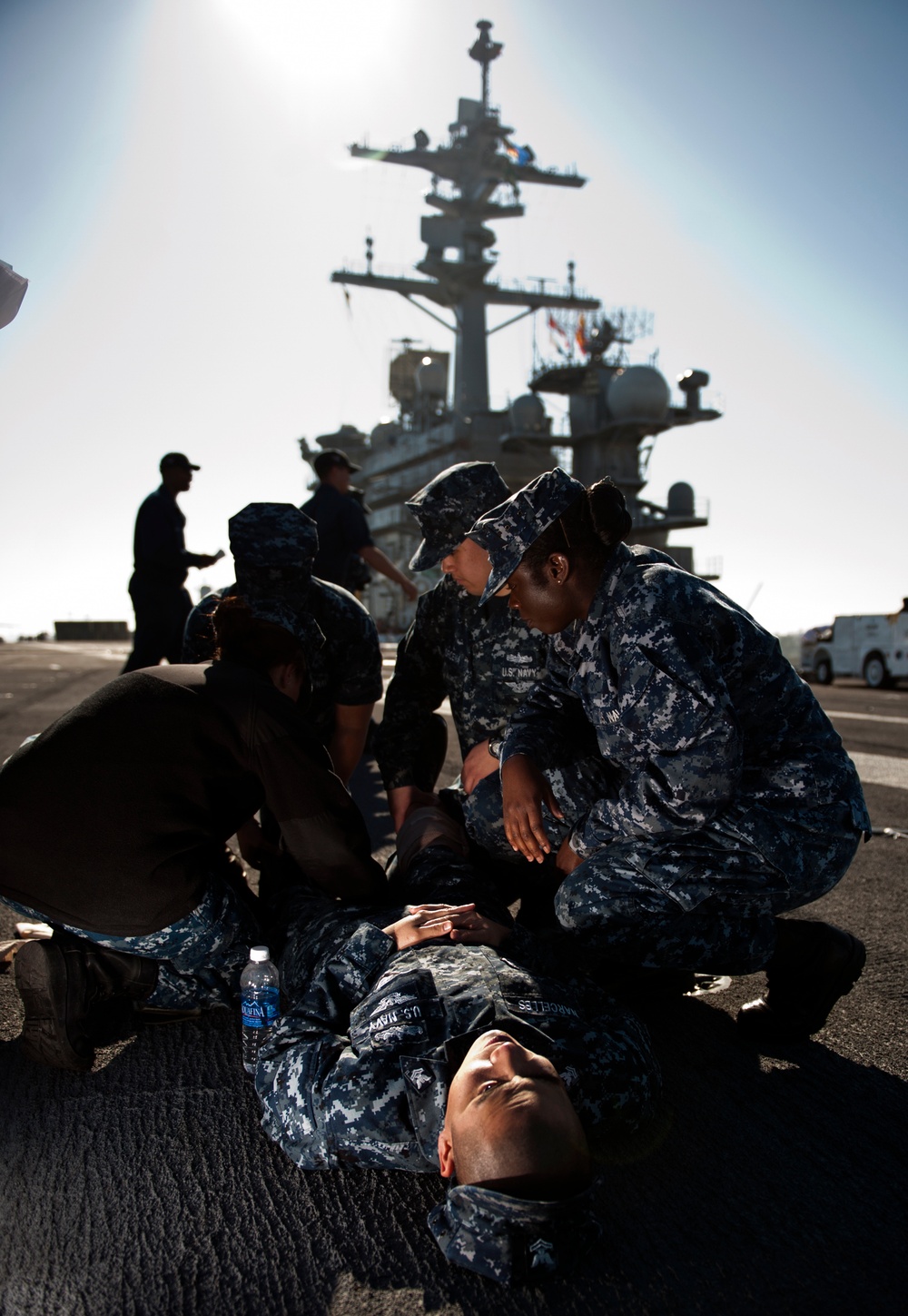 U.S. Navy personnel are conducting what appears to be a medical training drill or emergency response exercise on what looks like a pier or flight deck near a large naval vessel. Several sailors in Navy Working Uniform (NWU) digital camouflage are attending to a fellow sailor lying on the ground, with a water bottle placed nearby. A large warship, likely an aircraft carrier, is visible in the background.