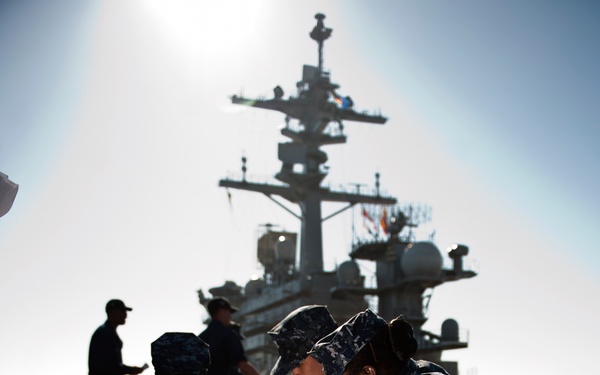 U.S. Navy personnel are conducting what appears to be a medical training drill or emergency response exercise on what looks like a pier or flight deck near a large naval vessel. Several sailors in Navy Working Uniform (NWU) digital camouflage are attending to a fellow sailor lying on the ground, with a water bottle placed nearby. A large warship, likely an aircraft carrier, is visible in the background.