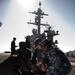 U.S. Navy personnel are conducting what appears to be a medical training drill or emergency response exercise on what looks like a pier or flight deck near a large naval vessel. Several sailors in Navy Working Uniform (NWU) digital camouflage are attending to a fellow sailor lying on the ground, with a water bottle placed nearby. A large warship, likely an aircraft carrier, is visible in the background.