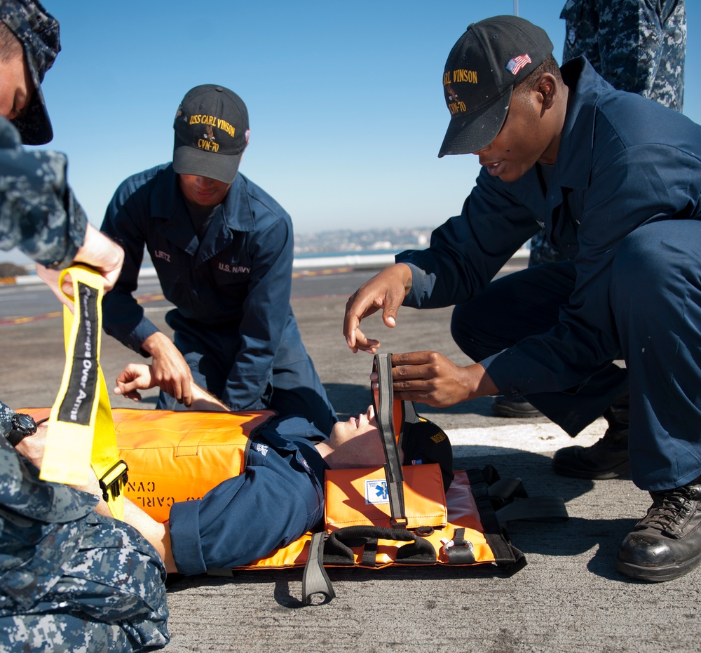 U.S. Navy sailors assigned to USS Carl Vinson (CVN-70) are conducting a medical casualty training exercise on what appears to be a flight deck or pier surface. Personnel are practicing securing a simulated casualty onto an orange stretcher/litter board. The exercise appears to be taking place in a sunny, outdoor environment near a waterfront.