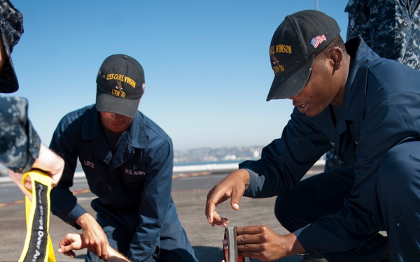 U.S. Navy sailors assigned to USS Carl Vinson (CVN-70) are conducting a medical casualty training exercise on what appears to be a flight deck or pier surface. Personnel are practicing securing a simulated casualty onto an orange stretcher/litter board. The exercise appears to be taking place in a sunny, outdoor environment near a waterfront.