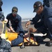 U.S. Navy sailors assigned to USS Carl Vinson (CVN-70) are conducting a medical casualty training exercise on what appears to be a flight deck or pier surface. Personnel are practicing securing a simulated casualty onto an orange stretcher/litter board. The exercise appears to be taking place in a sunny, outdoor environment near a waterfront.