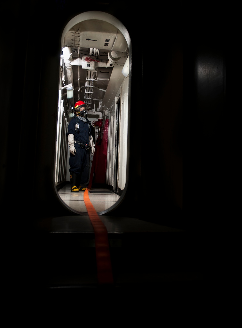 A person in protective gear stands in a narrow shipboard corridor viewed through an oval-shaped watertight door (hatch). The individual is wearing a dark blue coverall, red helmet, respiratory mask, white gloves, and yellow safety boots. An orange guideline or hose runs from the corridor through the hatch toward the camera.