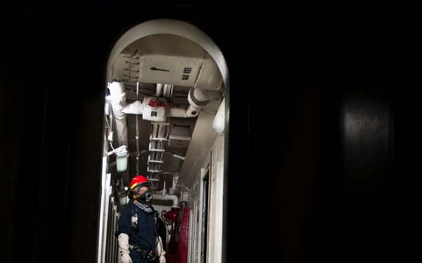 A person in protective gear stands in a narrow shipboard corridor viewed through an oval-shaped watertight door (hatch). The individual is wearing a dark blue coverall, red helmet, respiratory mask, white gloves, and yellow safety boots. An orange guideline or hose runs from the corridor through the hatch toward the camera.