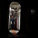 A person in protective gear stands in a narrow shipboard corridor viewed through an oval-shaped watertight door (hatch). The individual is wearing a dark blue coverall, red helmet, respiratory mask, white gloves, and yellow safety boots. An orange guideline or hose runs from the corridor through the hatch toward the camera.