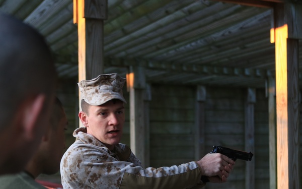 A military instructor in MARPAT camouflage uniform and cover demonstrates pistol handling technique at an indoor shooting range. He is addressing a small group of students visible in the foreground, holding a semi-automatic pistol in an extended grip position. The setting appears to be a covered firing range with wooden structural elements and a red shooting bench.