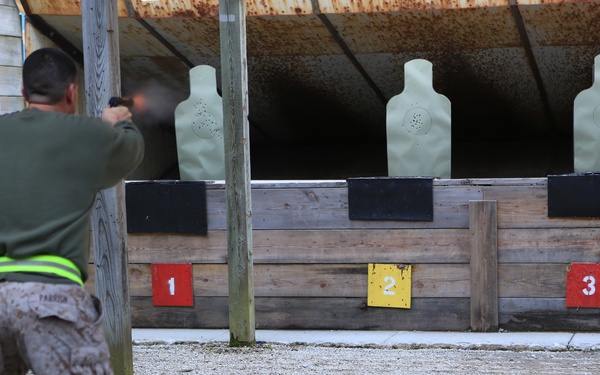 A military service member in camouflage utilities and a green sweatshirt is actively firing a pistol at a shooting range with three numbered silhouette targets. The shooter is positioned at lane 1, with muzzle flash visible at the firearm. The range features three numbered target positions (1, 2, 3) marked with colored placards and standard humanoid silhouette targets.