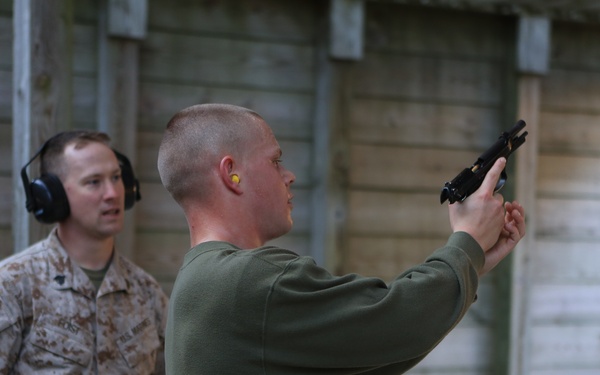 Two military personnel are at what appears to be an outdoor shooting range or training facility. One individual in a green sweatshirt and camouflage pants is aiming a pistol upward while the other, wearing full USMC MARPAT camouflage uniform with hearing protection, observes. The setting suggests a supervised firearms training exercise.