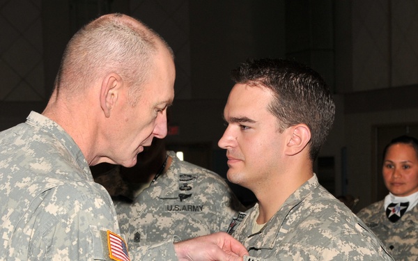 A U.S. Army officer is pinning a medal or award onto the uniform of a younger soldier during what appears to be a formal military ceremony. Both individuals are wearing Army Combat Uniforms (ACU) with digital camouflage pattern. Additional soldiers, including one wearing a 2nd Infantry Division patch and white gloves, are visible in the background.