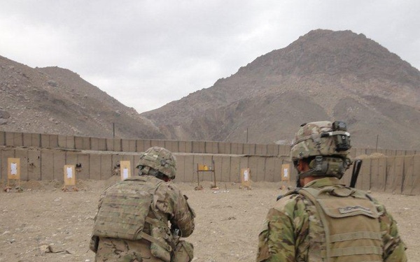 Two soldiers in full combat gear are walking toward a military firing range enclosed by HESCO barriers, set against an arid mountainous landscape. The range features multiple paper targets mounted on stands at varying distances. The terrain and environment are consistent with a forward operating base in a desert mountain region.