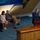 Hawaii State Medal of Honor ceremony pays tribute to fallen service members A formal ceremony or official meeting is taking place in what appears to be a legislative chamber or official hall. A military officer in a blue Air Force dress uniform is speaking at a wooden podium equipped with a microphone, while three civilians — two men and a woman — are seated in chairs on the stage listening attentively. A nameplate on the foreground podium reads 'DONNA MERCADO KIM, President,' and a gavel is visible on that podium.