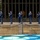 Hawaii State Medal of Honor ceremony pays tribute to fallen service members A ceremonial military honor guard, identifiable as U.S. Air Force personnel by their distinctive blue dress uniforms, is performing a drill or ceremony outside a large glass-facade building. The soldiers are holding rifles and standing in formation on a wet stone plaza, suggesting recent rainfall. A rope barrier separates the formation from the viewing area, and a decorative mosaic or tiled feature is visible in the foreground.