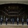 Hawaii State Medal of Honor ceremony pays tribute to fallen service members A military honor guard formation stands at attention in front of a large modern building with floor-to-ceiling tinted glass windows and bronze vertical mullions. Approximately eight uniformed personnel in dark blue dress uniforms are holding rifles, positioned in a evenly spaced line across the building's entrance. The scene appears to be a formal ceremonial drill or honor guard performance.