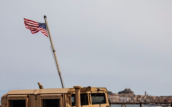 A tan/desert-colored U.S. military HMMWV (Humvee) is secured with chains on what appears to be the deck of a naval vessel. An American flag flies from a flagpole in the background, with a coastal city and hillside fortification visible across the water. The setting suggests a port call or amphibious operation in a Mediterranean location.