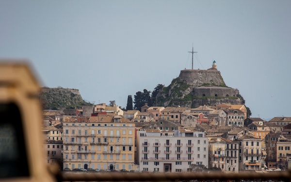 A coastal Mediterranean town is viewed from across a body of water, with a hilltop fortress crowned by a large cross dominating the skyline. The foreground contains a blurred vehicle hood and metal railing, suggesting the photo was taken from a ferry or boat deck. The architectural style, fortress structure, and church bell tower with a red dome are consistent with Corfu (Kerkyra), Greece.