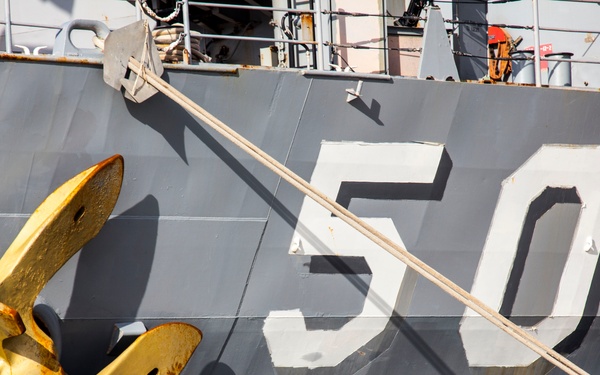 The image shows the hull of a naval warship marked with the hull number '50' in large white characters. A yellow-painted stockless anchor is visible on the port bow, secured in its hawsepipe. Mooring lines are visible crossing the frame diagonally, and deck railings with safety equipment are partially visible at the top.