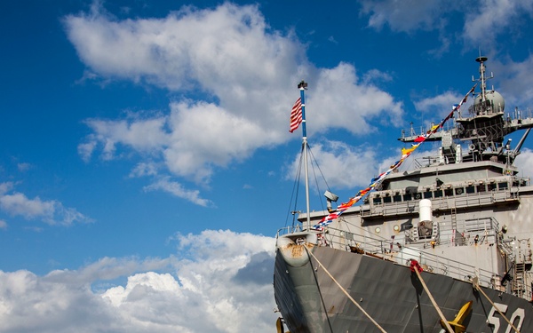 A large gray US Navy warship bearing hull number '50' is moored at a dock, viewed from a low bow-angle perspective. The vessel is decorated with colorful signal flags strung from its mast, and an American flag is visible flying near the superstructure. The background features calm blue water, a distant tree-covered landmass, and a partly cloudy sky.