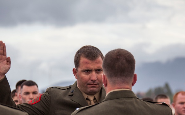 A group of U.S. Marines in dress green uniforms are participating in what appears to be a reenlistment or oath-of-office ceremony, with several individuals raising their right hands. The scene takes place outdoors against an overcast sky with mountains visible in the background. The central figure faces the camera with a serious expression while taking the oath.