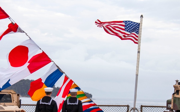 Two U.S. Navy sailors in dress blues stand at attention on the deck of a naval vessel, surrounded by military vehicles and a display of international signal flags. The American flag flies prominently from a mast, while a large bundle of nautical signal flags — including the Japanese flag — streams in the wind. The scene appears to depict a formal naval ceremony or port visit, likely in Japan based on contextual clues.