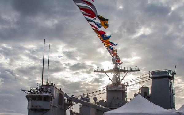 A formal military ceremony is taking place on the deck of a naval warship, likely a commissioning or decommissioning event. The deck is decorated with a string of international signal flags (rainbow bunting) stretching diagonally across the sky, and a white event tent has been erected on the deck. Naval personnel in dress uniforms are present alongside what appear to be color guard members holding flags.