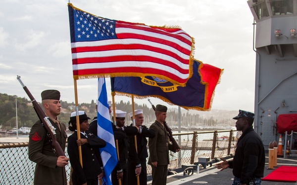 A military color guard ceremony is taking place on the deck of a U.S. Navy vessel. Personnel in various uniforms, including U.S. Marines and Navy sailors, are holding flags including the U.S. flag, U.S. Navy flag, and what appears to be the Greek flag. A sailor in camouflage utilities is approaching the color guard formation, likely conducting an inspection or directing the ceremony.