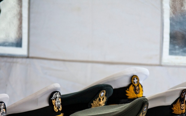 Several military and naval officer dress caps are displayed on a white cloth-covered table. The caps vary in color and style, including white naval caps with gold embroidery and black brims, dark green military caps, and one olive/field-style cap. A uniformed individual is partially visible on the left side of the frame.
