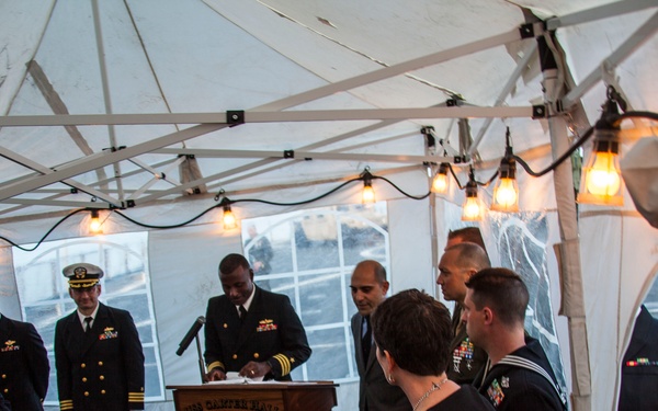 A formal military ceremony is taking place under a white tent, with naval officers and civilians gathered around a podium. The podium bears a placard reading 'USS CARTER HALL (LSD 50)', indicating this is an official U.S. Navy event aboard or associated with that ship. String lights illuminate the tent as a senior naval officer addresses or conducts proceedings at the lectern.
