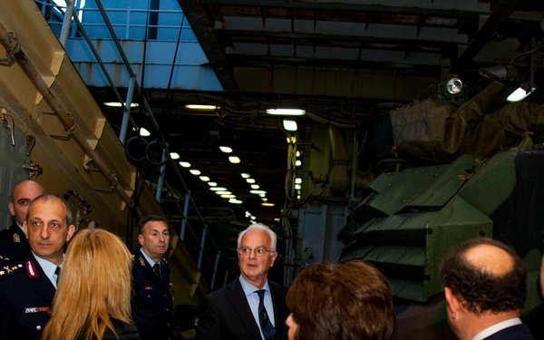 A group of military officers and civilian officials are gathered inside what appears to be a military hangar or vehicle storage facility. The group includes uniformed personnel and civilians in formal attire, seemingly engaged in a briefing or tour. Large military vehicles or equipment are visible on both sides of the gathering.