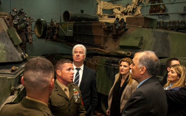 A group of civilians and military personnel are gathered in what appears to be a military vehicle storage facility or motor pool. Two uniformed Marines are briefing or conversing with a group of approximately five civilians dressed in business attire. The setting is flanked by large military armored vehicles, likely M1 Abrams tanks, in camouflage paint.