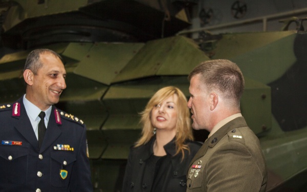 Two military officers in different uniforms are engaged in conversation, with a civilian woman standing between them. The setting appears to be inside a military facility or museum, with a camouflage-painted armored vehicle visible in the background. The interaction appears formal and cordial, suggesting a diplomatic or official military meeting.