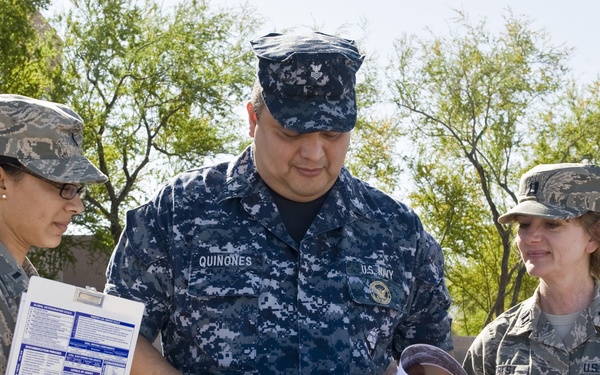 Three military personnel are gathered outdoors reviewing documents on clipboards. The group includes one U.S. Navy sailor and two U.S. Air Force airmen, suggesting an inter-service coordination activity. They appear to be at a military installation, likely conducting an inspection, assessment, or administrative review.