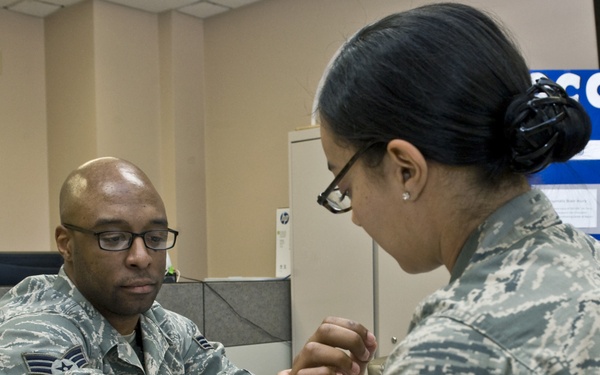 Two U.S. Air Force personnel in ABU (Airman Battle Uniform) camouflage are seated at a table where one airman appears to be receiving occupational therapy or a hand/wrist examination. A blue towel is placed under the patient's arm on the table, suggesting a clinical or therapeutic setting. A blue banner partially visible in the background reads 'OCCUPA... THE...' consistent with an Occupational Therapy display.