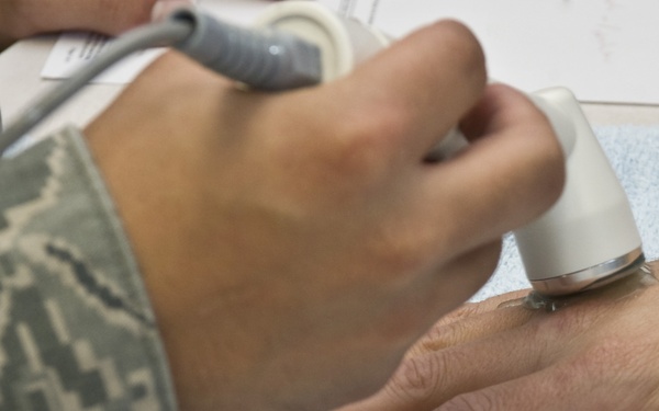 A medical professional wearing US Air Force camouflage (ABU pattern) uniform is applying an ultrasound probe to the back of a patient's hand. The hand rests on a light blue medical towel, and the ultrasound transducer with attached cable is being pressed against the skin. This appears to be a clinical or medical examination setting, likely on a military base.